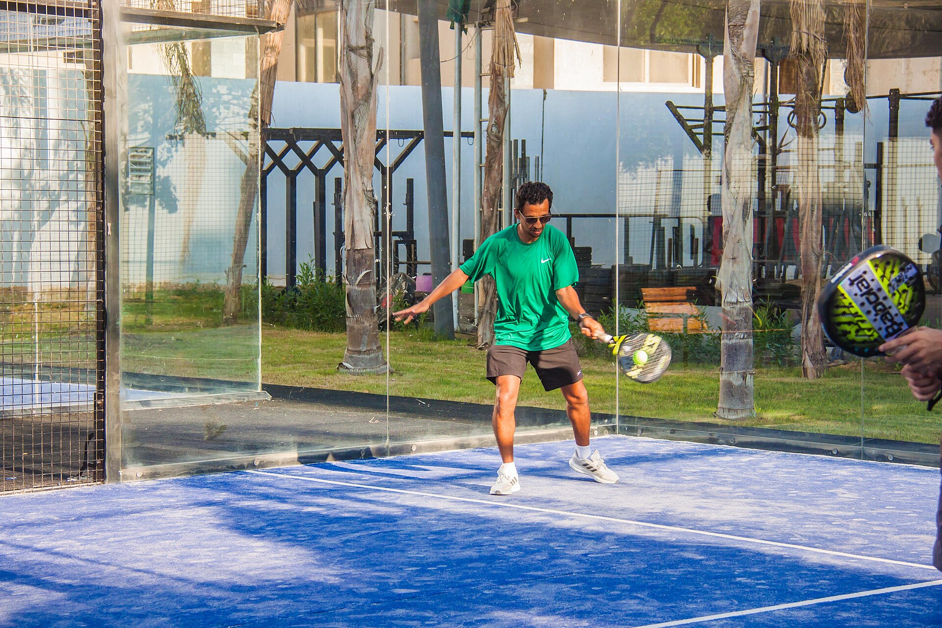 Pemain padel memukul bola forehand di lapangan sintetis biru berdinding kaca saat laga ganda