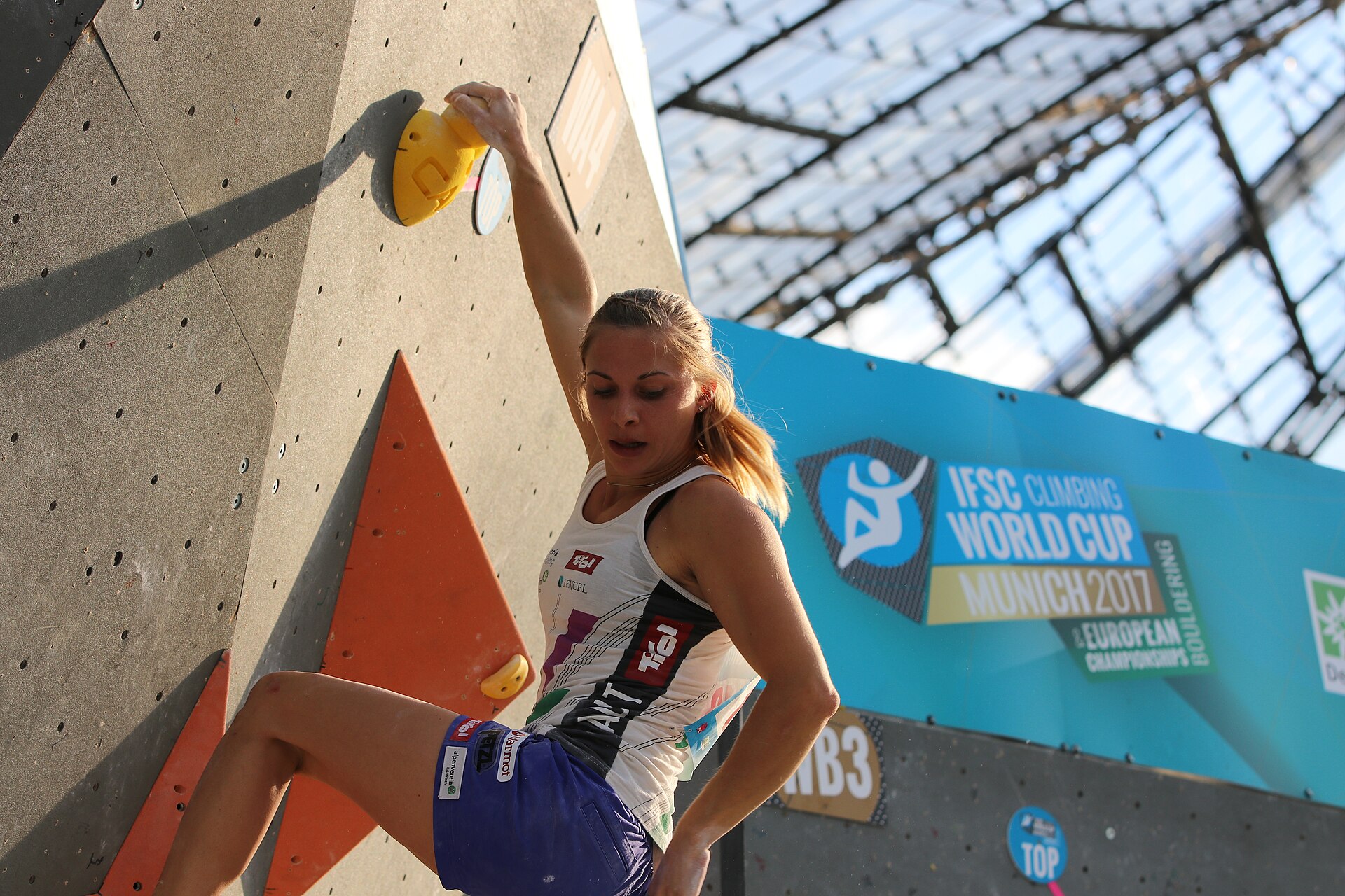 Atlet panjat tebing meraih pegangan kuning di dinding boulder indoor dengan spanduk IFSC Climbing World Cup di latar belakang