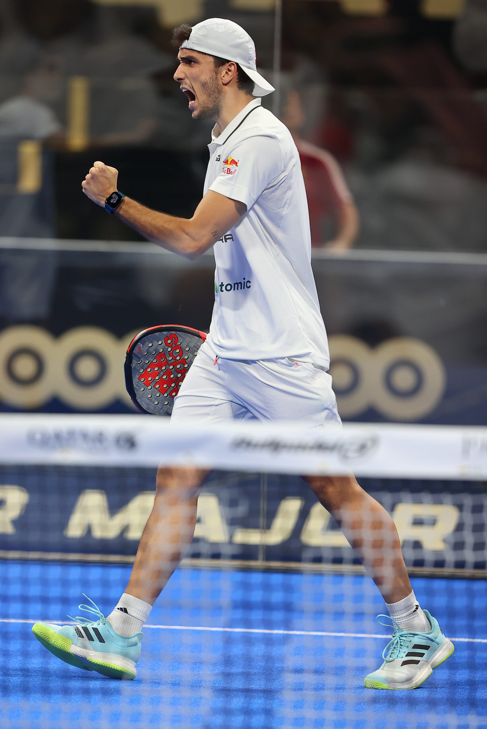Pemain padel profesional mengepalkan tangan dan berteriak merayakan poin di depan net, mengenakan jersey bersponsor Red Bull dan Adidas di ajang Premier Padel Major.