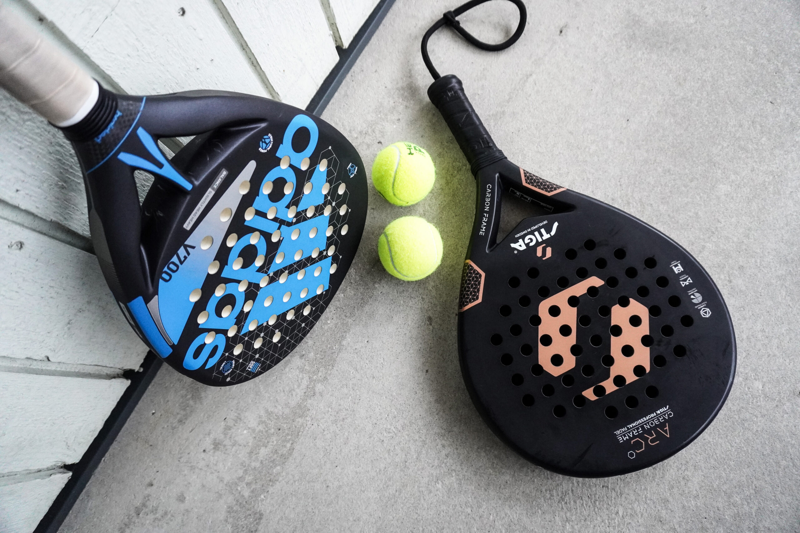 Dua raket padel karbon dan dua bola kuning tergeletak berdampingan di lantai kayu.