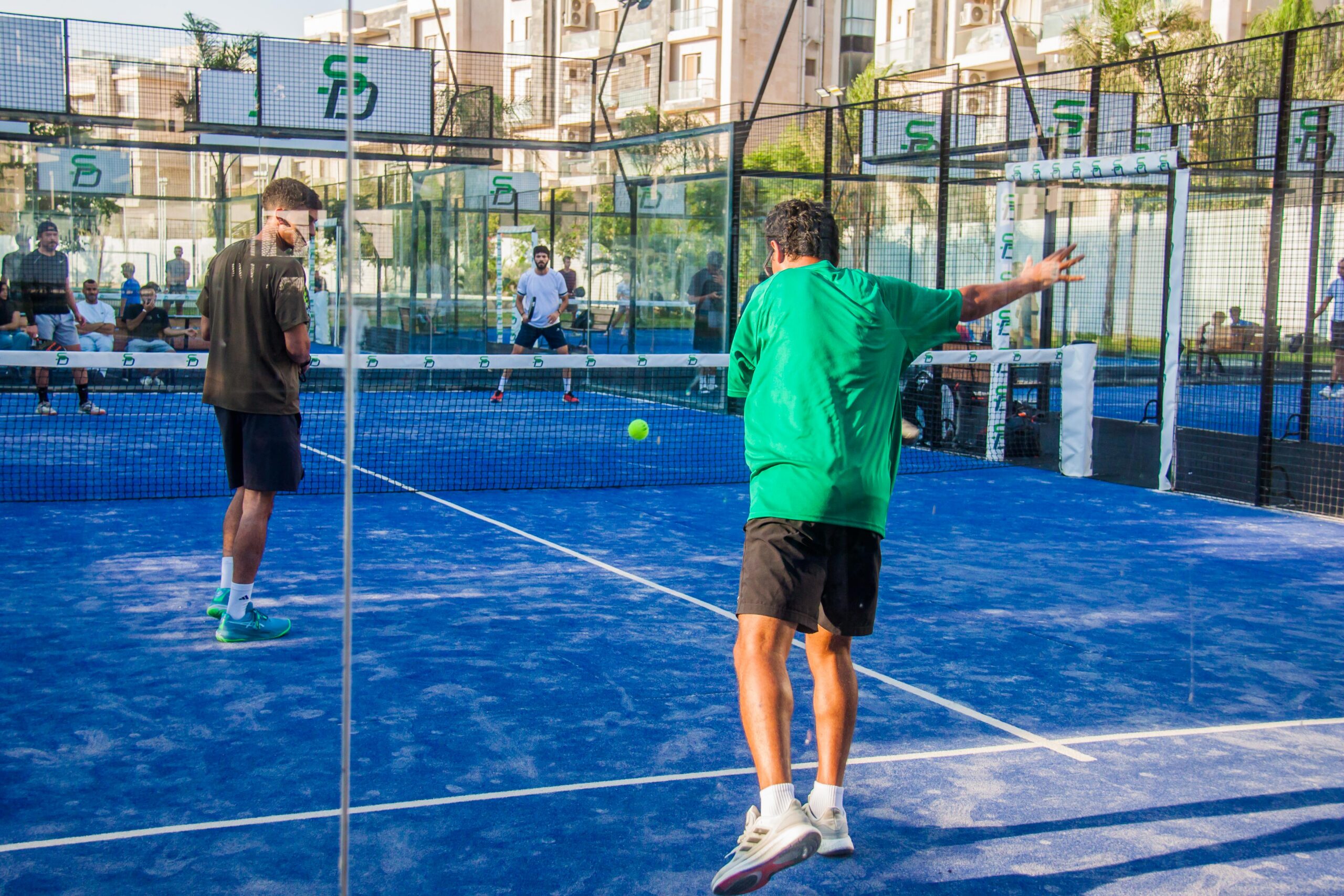 Pemain padel melompat melakukan smes dalam pertandingan ganda di lapangan klub outdoor, disaksikan pemain lain yang menunggu giliran dengan gedung apartemen di latar belakang.