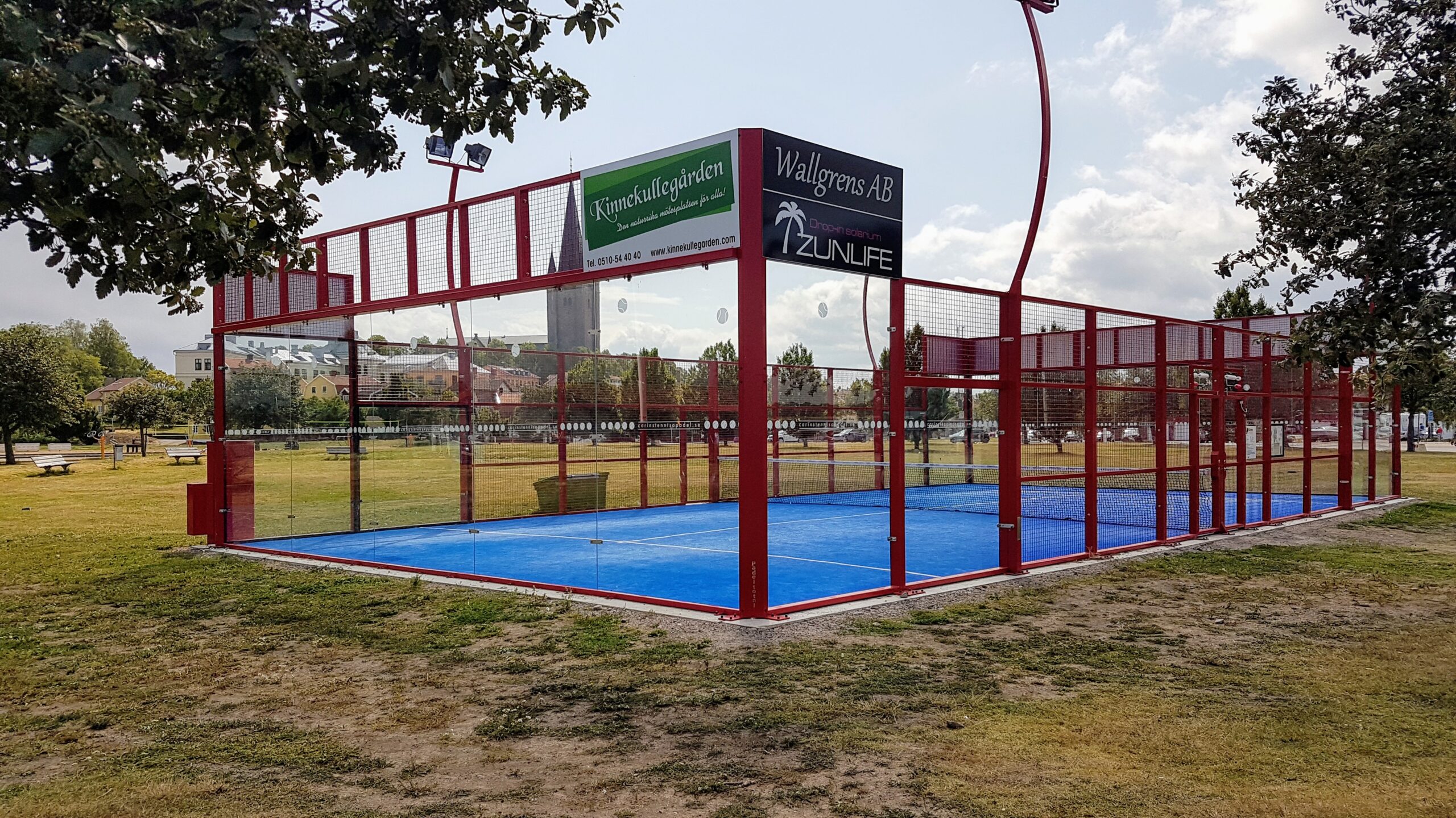 Lapangan padel kosong dikelilingi rumput taman dan pepohonan pada sore yang tenang.