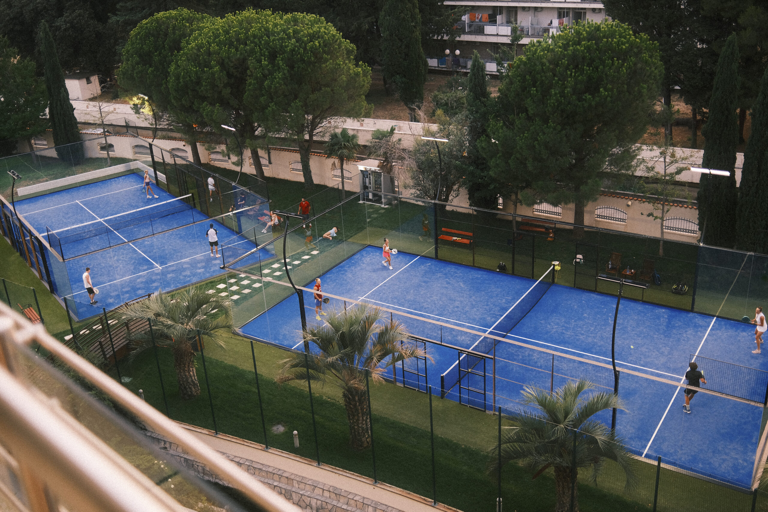 Pemandangan dari atas beberapa lapangan padel biru dengan pemain ganda dikelilingi pohon pinus dan palem.