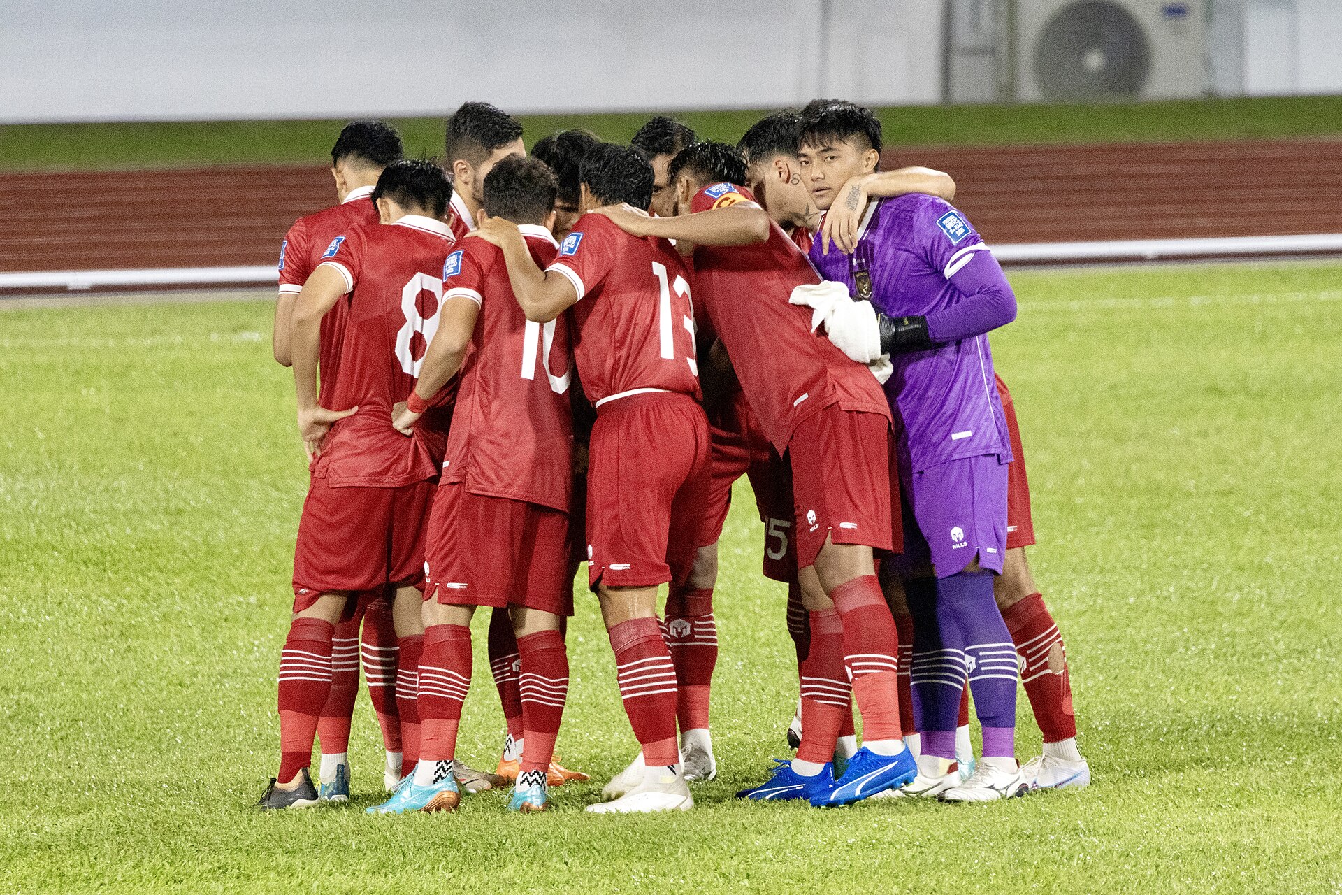 Pemain Timnas Indonesia berseragam merah berkumpul membentuk lingkaran di lapangan sebelum pertandingan, kiper berseragam ungu merangkul rekan setim
