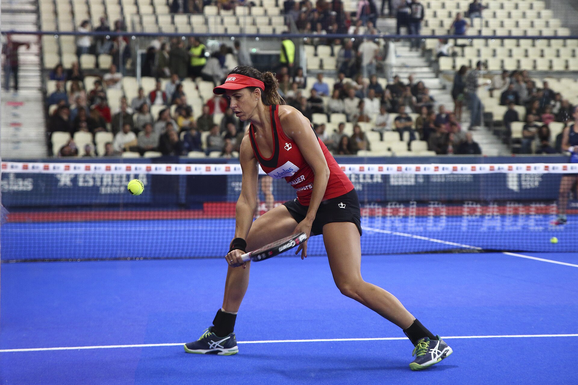 Ilustrasi: pemain padel putri berbaju merah dengan visor menjangkau bola rendah saat laga turnamen profesional di lapangan berkaca