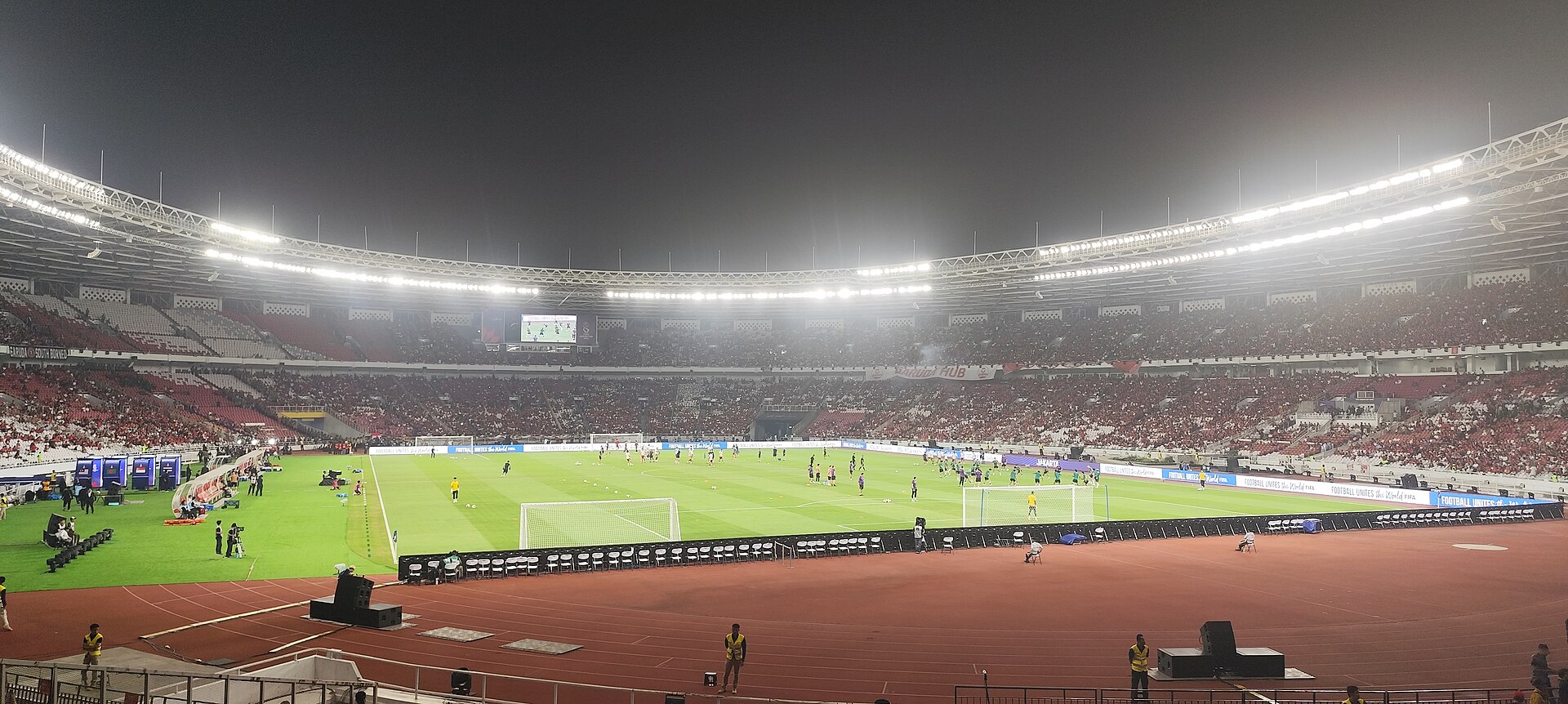 Suasana Stadion Utama Gelora Bung Karno, Jakarta, pada malam hari dilihat dari tribun: lampu sorot menerangi lapangan tempat para pemain pemanasan, sementara tribun dipenuhi suporter berkostum merah.