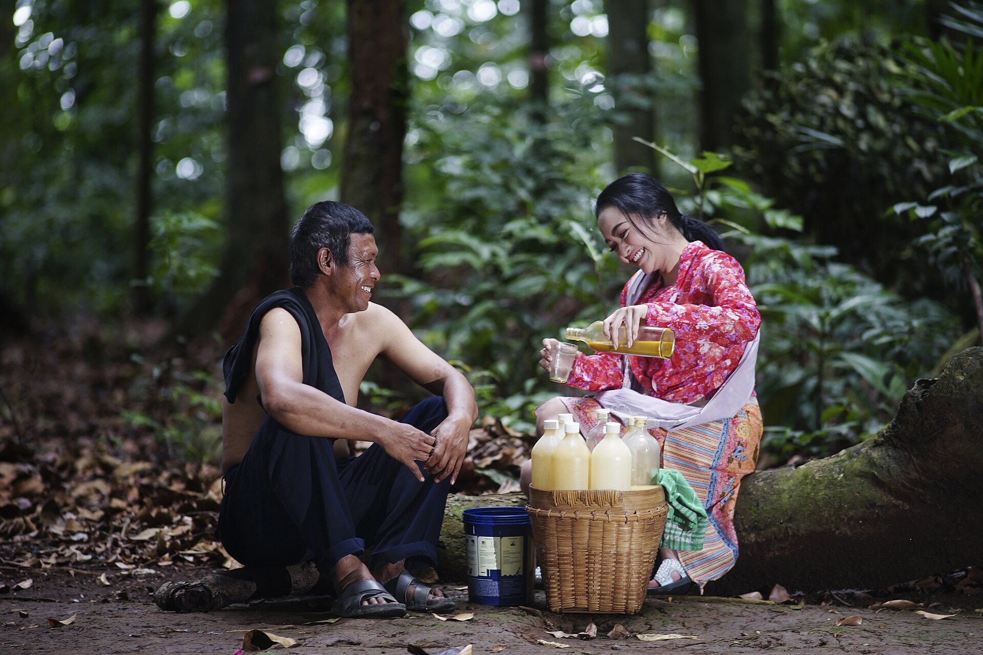 Seorang perempuan berkebaya menuang jamu dari botol ke gelas, dengan bakul bambu berisi botol jamu di depannya, di tengah hutan