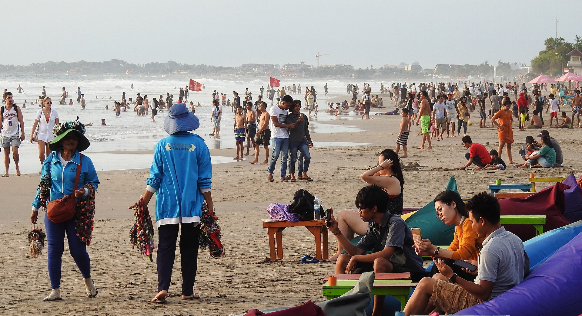 Pantai Legian di Bali dipadati pengunjung saat sore: pedagang asongan berjalan di pasir dan banyak orang bermain di tepi ombak