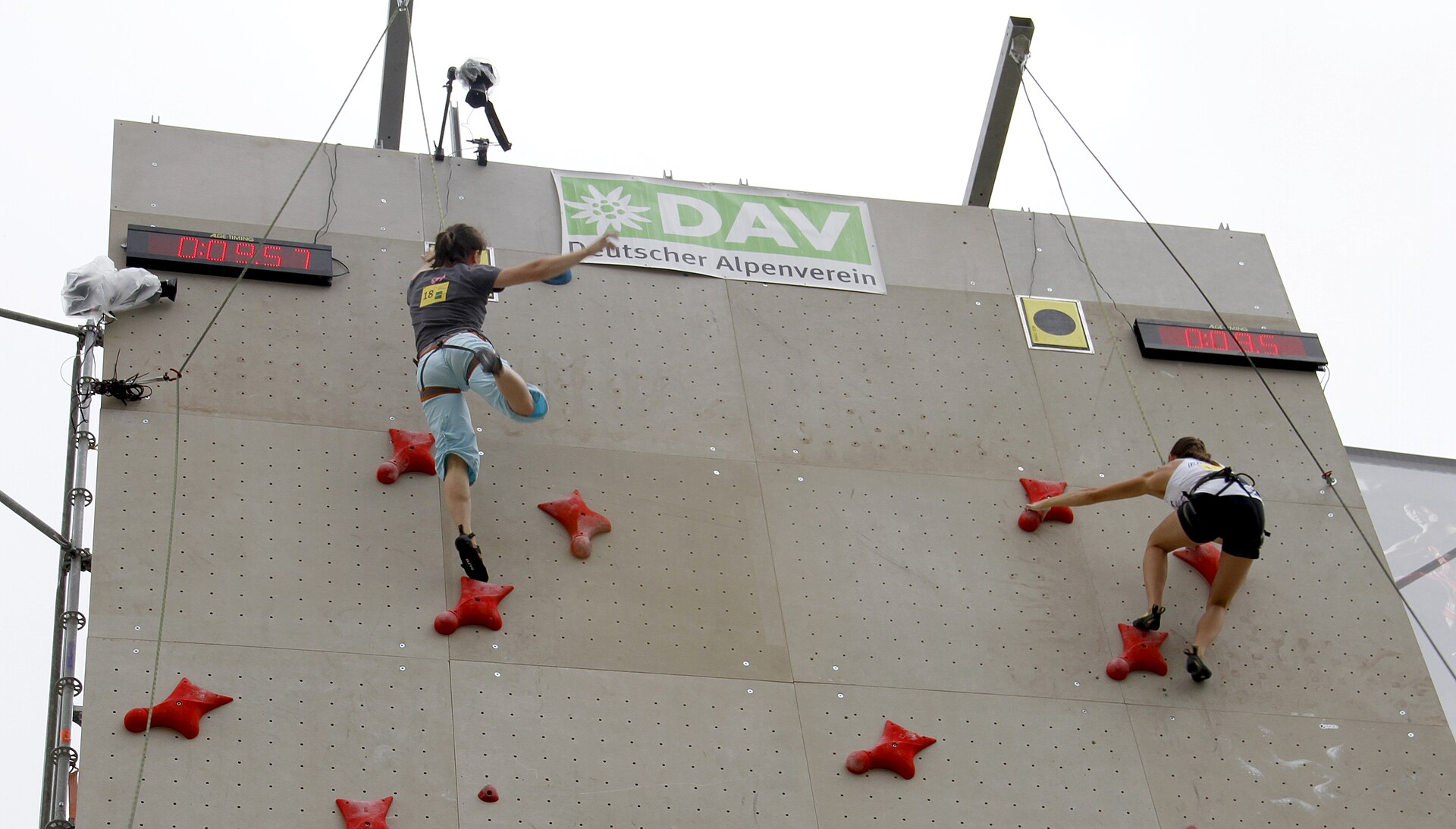 Dua atlet speed climbing memanjat berdampingan di dinding tinggi dengan papan waktu, foto ilustrasi arena adu cepat.