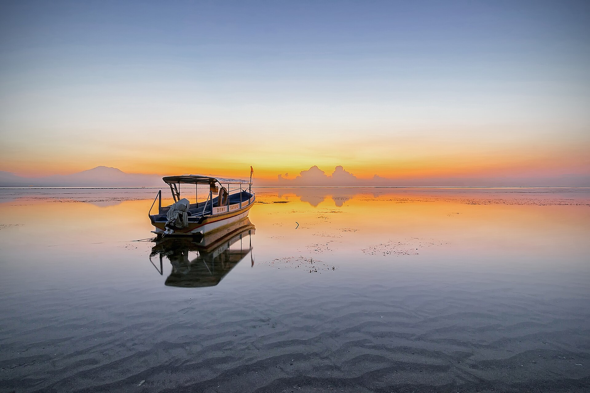 Perahu wisata bersandar di perairan dangkal Pantai Sanur saat matahari terbit, dengan langit jingga terpantul di air tenang