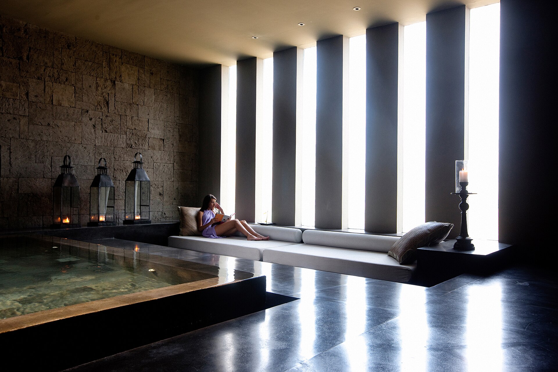 Ruang relaksasi spa di sebuah resor Bali dengan kolam rendam, lentera berlilin, dan seorang perempuan membaca di dipan
