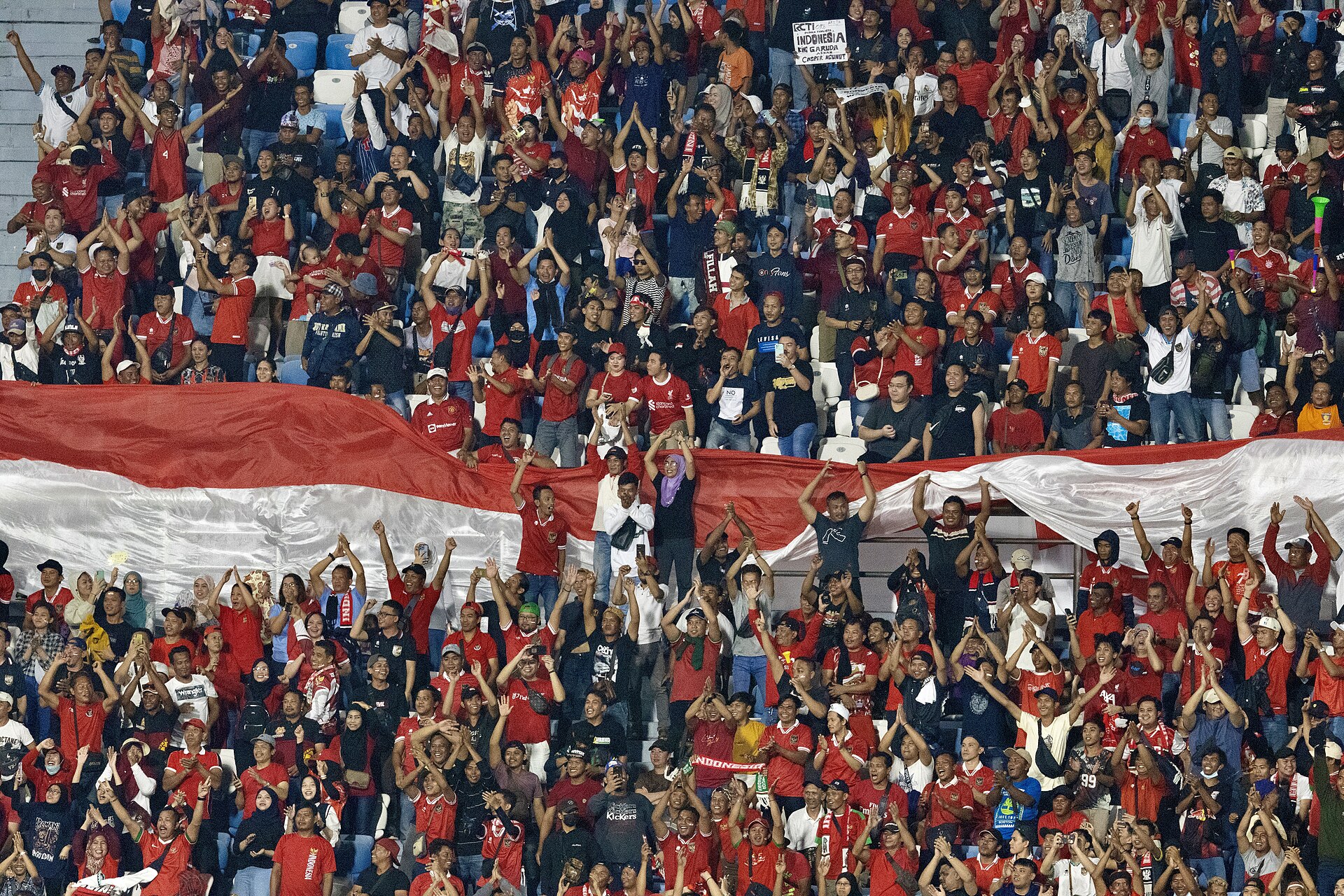 Suporter Timnas Indonesia berkostum merah bersorak dan bertepuk tangan di tribun, dengan bendera Merah Putih raksasa terbentang di antara kerumunan.