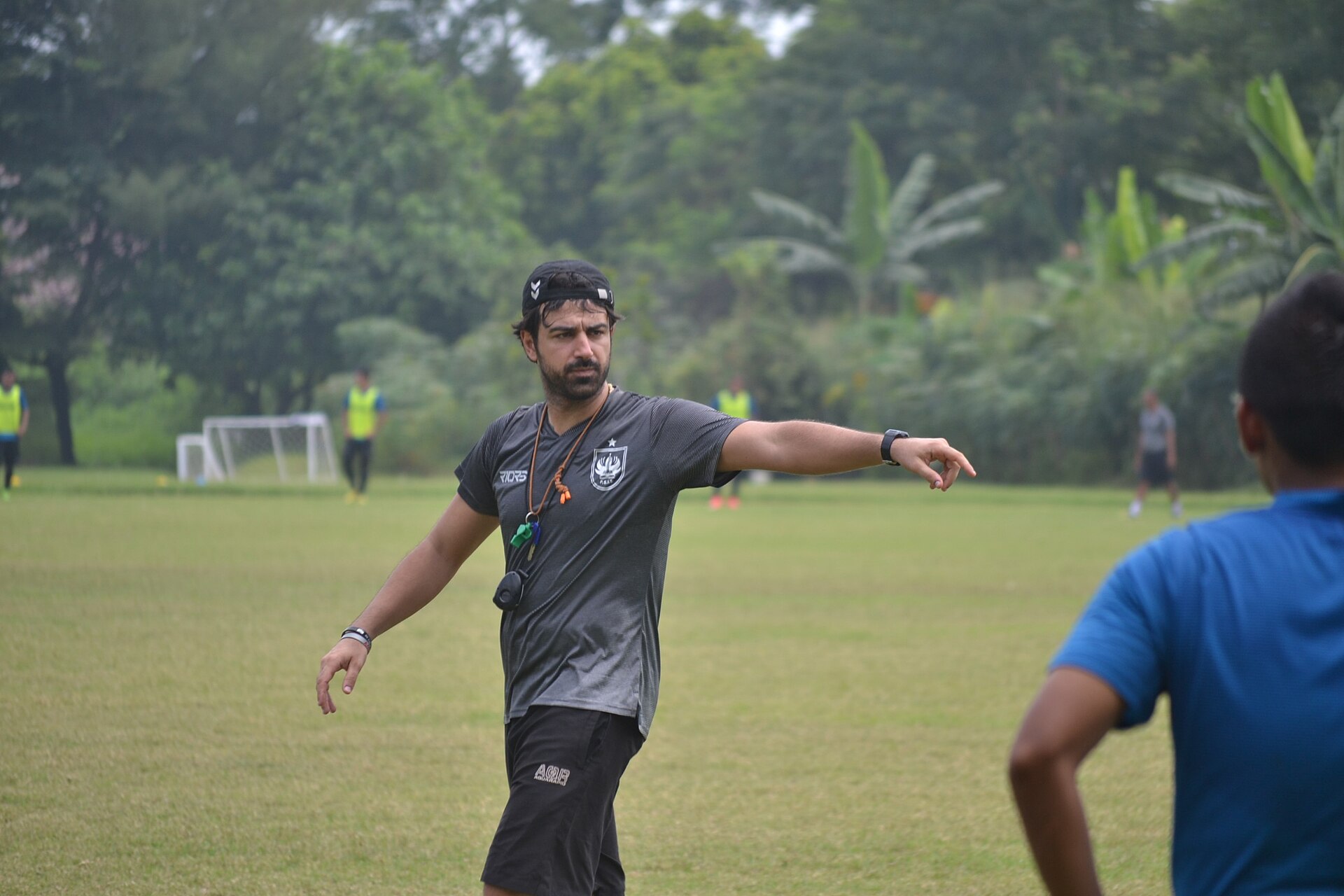 Seorang pelatih berkaus abu-abu klub PSIS Semarang menunjuk dan memberi arahan dalam sesi latihan di lapangan terbuka, dengan pemain berompi kuning dan sebuah gawang di belakangnya.
