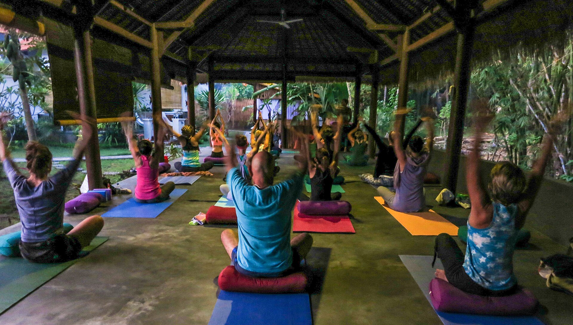 Peserta kelas yoga pagi mengangkat tangan di atas matras dalam bale terbuka beratap alang-alang di Amed, Bali
